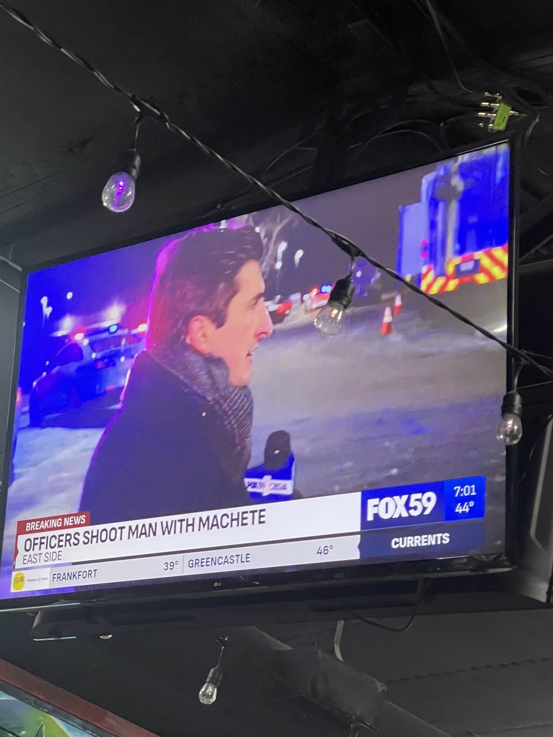 A TV screen displays a news report about an officer-involved shooting incident, with a reporter on the scene. “Officers shoot man with machete”